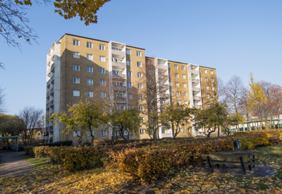 Ett flerfamiljshus i ljusgul tegel med åtta våningar och vita balkonger, omgivet av ett höstlandskap. Löv har fallit på marken och några träd har gula och orangefärgade blad. I förgrunden finns en liten park med buskar, träd och en bänk. Himlen är klarblå och solen lyser, vilket skapar en lugn och fridfull känsla.