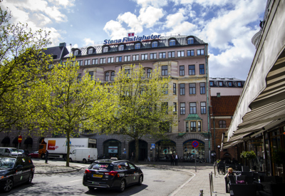 Stortorget i Malmö med bilar som kör och står parkerade framför en stor byggnad med skylten "Stena Fastigheter" på taket. Längs torget står gröna vårträd och till höger syns en uteservering med människor. Himlen är delvis molnig men ljus.
