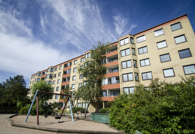 Flerfamiljshus i ljust tegel med röd takkant, omgiven av grönska och en lekplats med gungställning i förgrunden under en klarblå himmel.