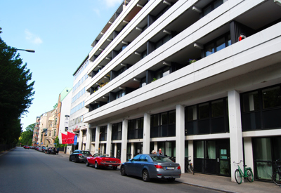 Byggnaden Väveriet i Malmö, med en modern fasad i vitt och svart och breda balkonger längs hela husets längd. Flera bilar är parkerade längs gatan, där även några cyklar står lutade mot fasaden. Till vänster i bild syns träd och äldre byggnader längre ner på gatan under en klarblå himmel.