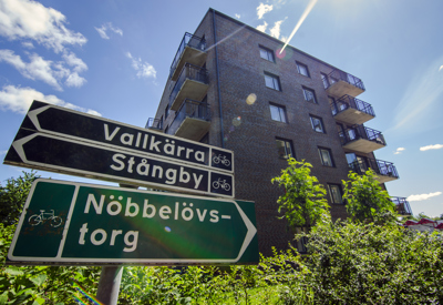 Cykelvägsskyltar som pekar mot Vallkärra, Stångby och Nöbbelövstorg, med en femvåningsbyggnad i mörkt tegel i bakgrunden. Byggnaden har flera balkonger, och runt omkring finns grönska. Soligt väder med blå himmel och några moln.