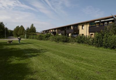 En låg bostadsbyggnad med gula tegelväggar och mörka trädetaljer löper längs höger sida av bilden. Byggnaden har balkonger och är omgiven av en tät häck. I förgrunden breder en välklippt gräsmatta ut sig, och till vänster promenerar en person med en hund. Himlen är delvis molnig med strimmor av blått, vilket ger en lugn och behaglig stämning i området.