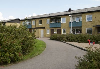 En tvåvånings bostadsbyggnad i gult tegel med balkonger målade i blått. Byggnaden omges av buskar och en asfalterad gångväg leder förbi husets entréer. Till höger syns en liten lekplats med sand och ett rött fjädergungdjur. Det är en ljus dag med blå himmel och några moln, vilket bidrar till en trivsam atmosfär i bostadsområdet.