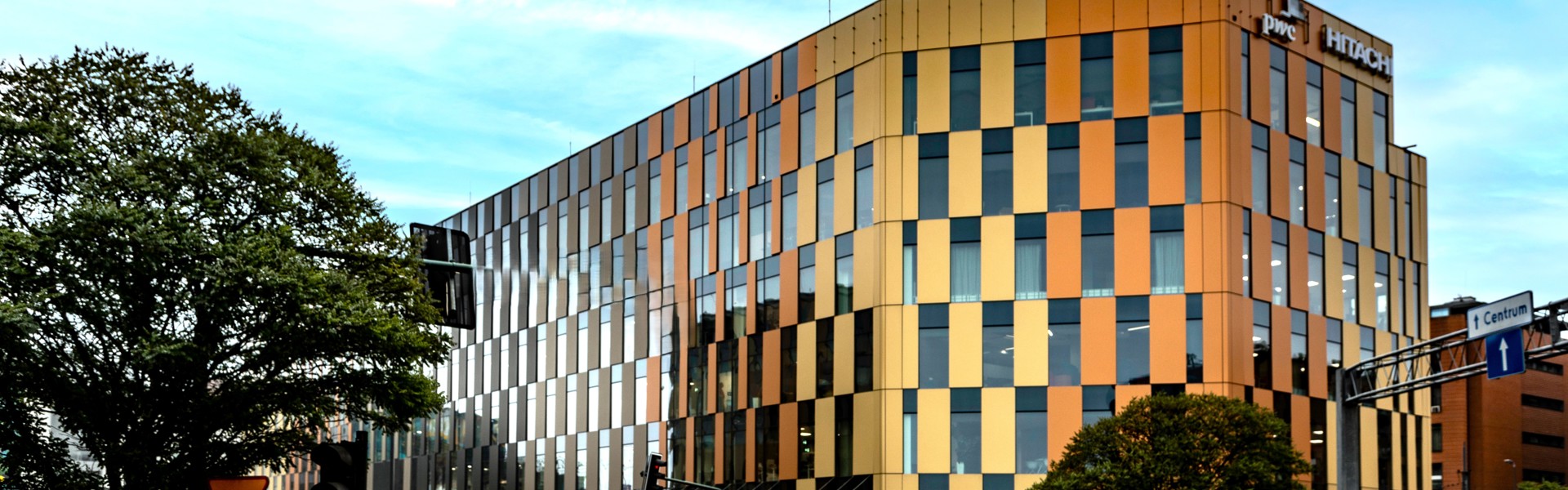 A modern office building with a golden and glass facade, featuring vertical panels of varying shades. The building is surrounded by trees and urban infrastructure, with a bright blue sky in the background