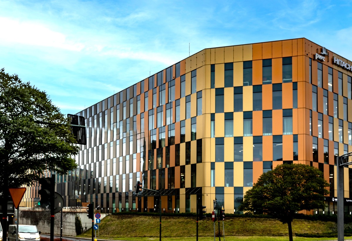 A modern office building with a golden and glass facade, featuring vertical panels of varying shades. The building is surrounded by trees and urban infrastructure, with a bright blue sky in the background