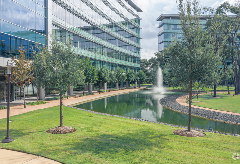 Landscaped office park with modern glass buildings, a curved pond with a fountain, walking paths, and rows of young trees.
