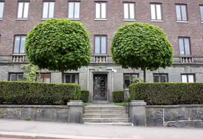 Entré till ett äldre stenhus, omgiven av välskötta häckar och träd, med en stenlagd gångstig som leder fram till en dekorativ dörr.
