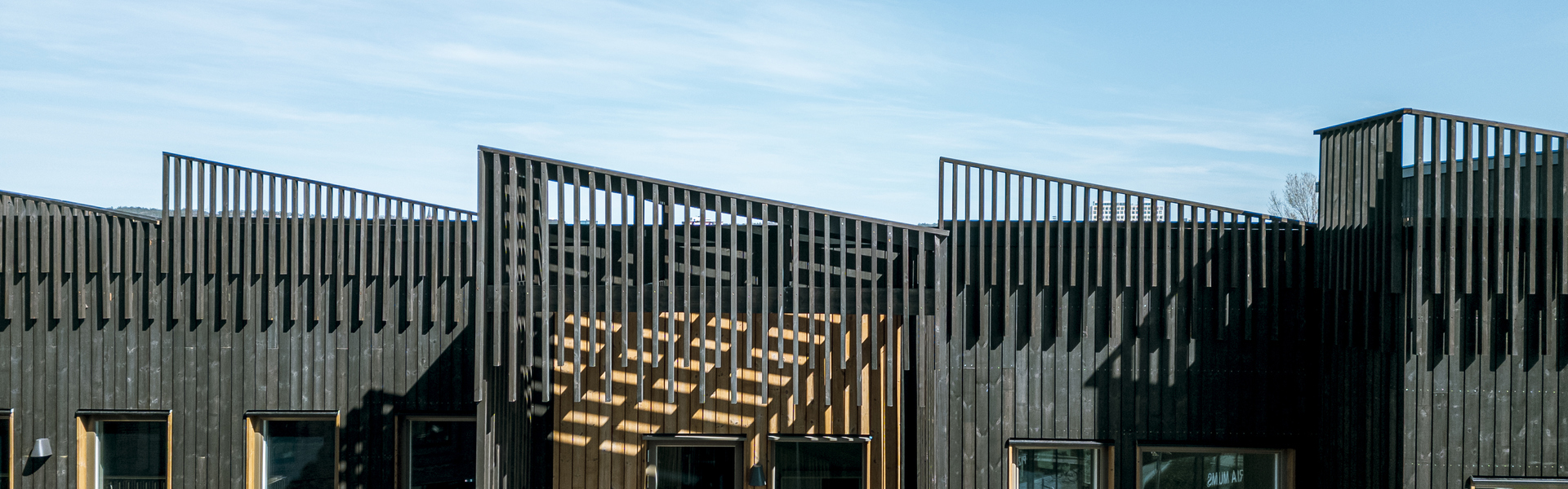 Fasad på ett modernt flerbostadshus i mörkt trä med balkonger, takterrasser och stora fönster under en klarblå himmel.