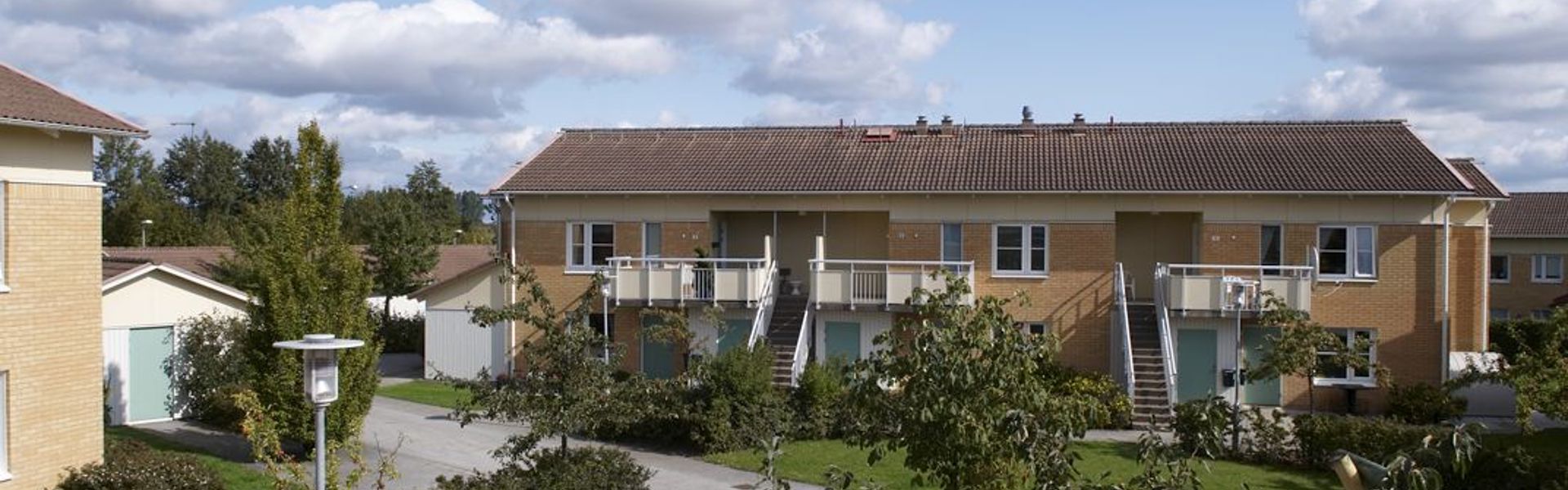 En solig dag i ett bostadsområde med tvåvåningshus i tegel. Husen har balkonger och omges av grönska och träd. En lekplats med sandlåda och gungställning syns i förgrunden. Moln pryder den blå himlen.