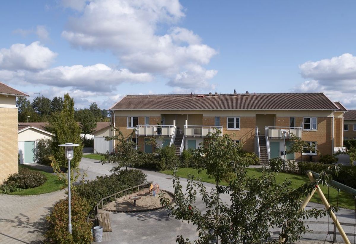 En solig dag i ett bostadsområde med tvåvåningshus i tegel. Husen har balkonger och omges av grönska och träd. En lekplats med sandlåda och gungställning syns i förgrunden. Moln pryder den blå himlen.