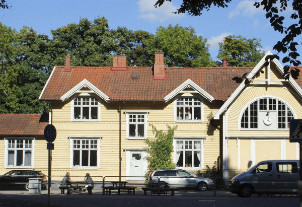 Ett gult trähus med rött tegeltak och vita fönster, fotograferat i dagsljus med grönskande träd i bakgrunden. Framför huset står några bilar parkerade och två personer sitter på en bänk vid trottoaren.