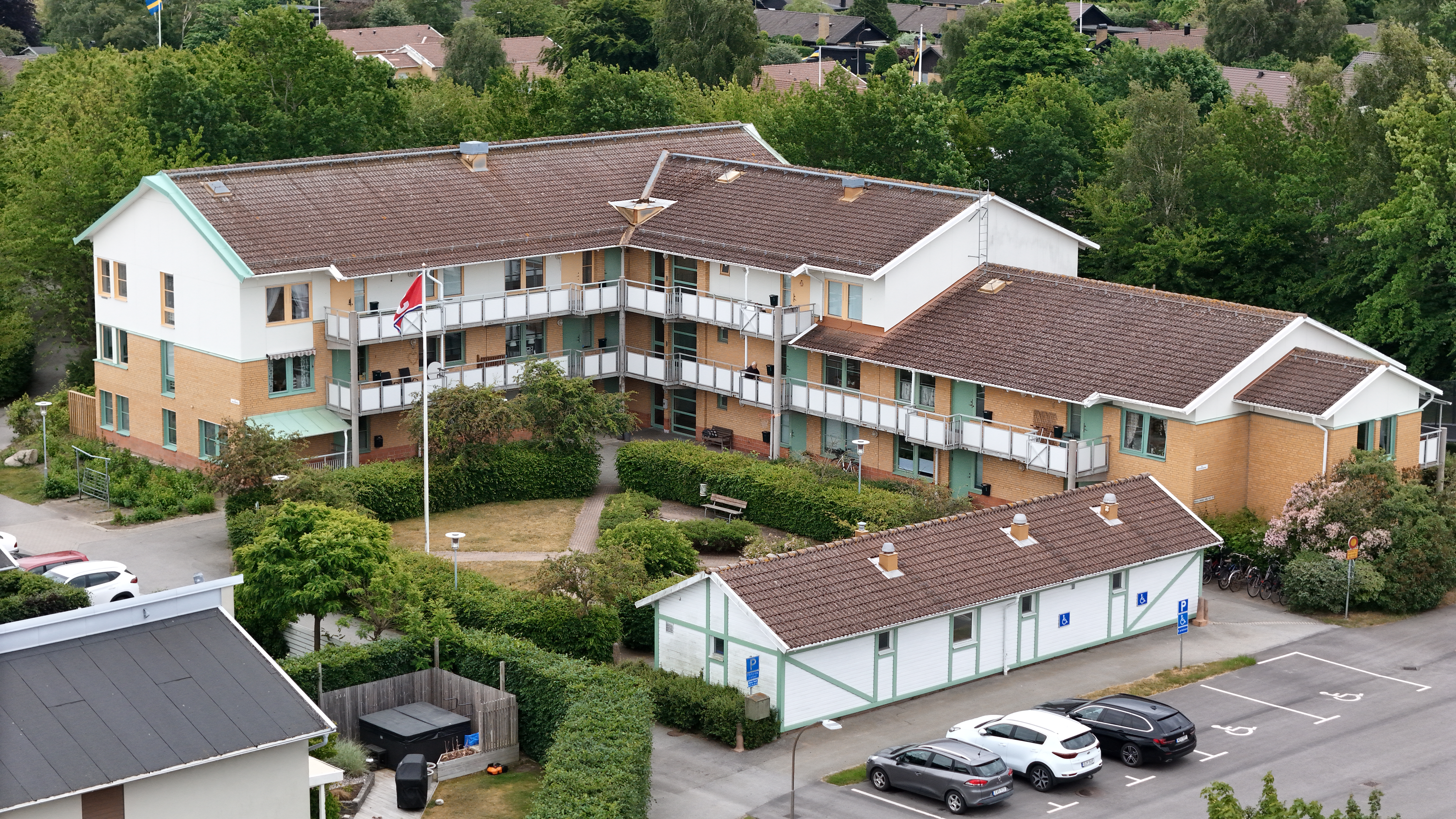Flygbild över ett lågt bostadshus med balkonger, innergård, grönska och parkeringsplatser intill.