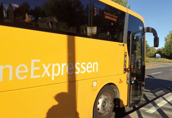 En gul buss från SkåneExpressen står stilla vid en hållplats en solig dag. Bussens dörr är öppen och en skugga från en person syns på sidan av bussen. Destinationen på skylten visar 'Simrishamn'.