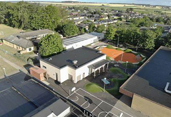 Flygfoto över en skolgård med lekplatser, basketplan och skolbyggnader omgivna av träd och bostadshus. I bakgrunden syns öppna fält och ett samhälle med småhus.
