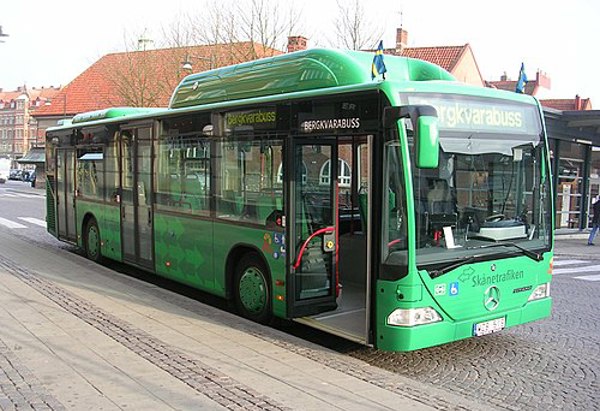 En grön stadsbuss från Skånetrafiken står vid en busshållplats. Bussen har texten 'Bergkvarabuss' ovanför dörrarna och är parkerad längs en trottoar med stenbeläggning. Det syns några hus med tegeltak och träd utan löv i bakgrunden.