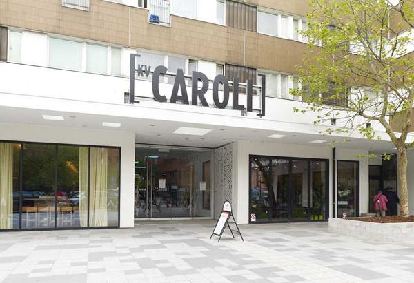 Entrén till byggnaden 'Kv Caroli' med stora glasdörrar och fönsterpartier. Ovanför entrén sitter en tydlig skylt med texten 'KV CAROLI'. Framför ingången finns ett öppet torg med ljusgrå markplattor i olika nyanser, ett träd planterat i en upphöjd låda och en skyltställning. I bakgrunden syns reflektioner av grönska och omkringliggande byggnader i glaset.