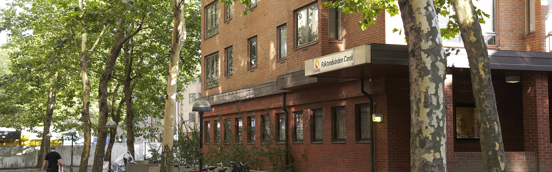 Ett tegelrött flerbostadshus med en skylt som visar 'Folktandvården Caroli' vid entrén. Byggnaden är omgiven av träd och cykelställ. En person går längs en stenlagd gångväg till vänster i bilden.