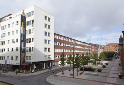 Ett gathörn med ett vitt flerbostadshus med butik och uteservering i bottenplan. Intill löper en bred gång- och cykelväg kantad av träd och planteringar. Flerbostadshus i röd tegelstil sträcker sig längs gatan under en delvis molnig himmel.