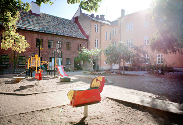 En solbelyst innergård med en lekplats som innehåller rutschkana, klätterställning och fjädergungor i färgglada färger. Gården är omgiven av äldre flerbostadshus i tegel med stora fönster. Träd och skuggor från lövverket ramar in platsen, och solens strålar skapar en varm, inbjudande atmosfär.
