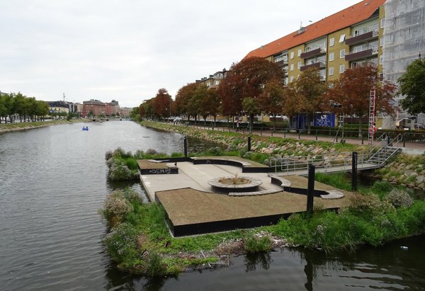 En flytande plattform i en kanal med sittplatser och växtlighet, omgiven av vatten och gröna växter. I bakgrunden syns en allé med höstfärgade träd längs en gångväg, samt flerbostadshus med röda tak och balkonger. Till vänster syns delar av en stadsmiljö under en molnig himmel.