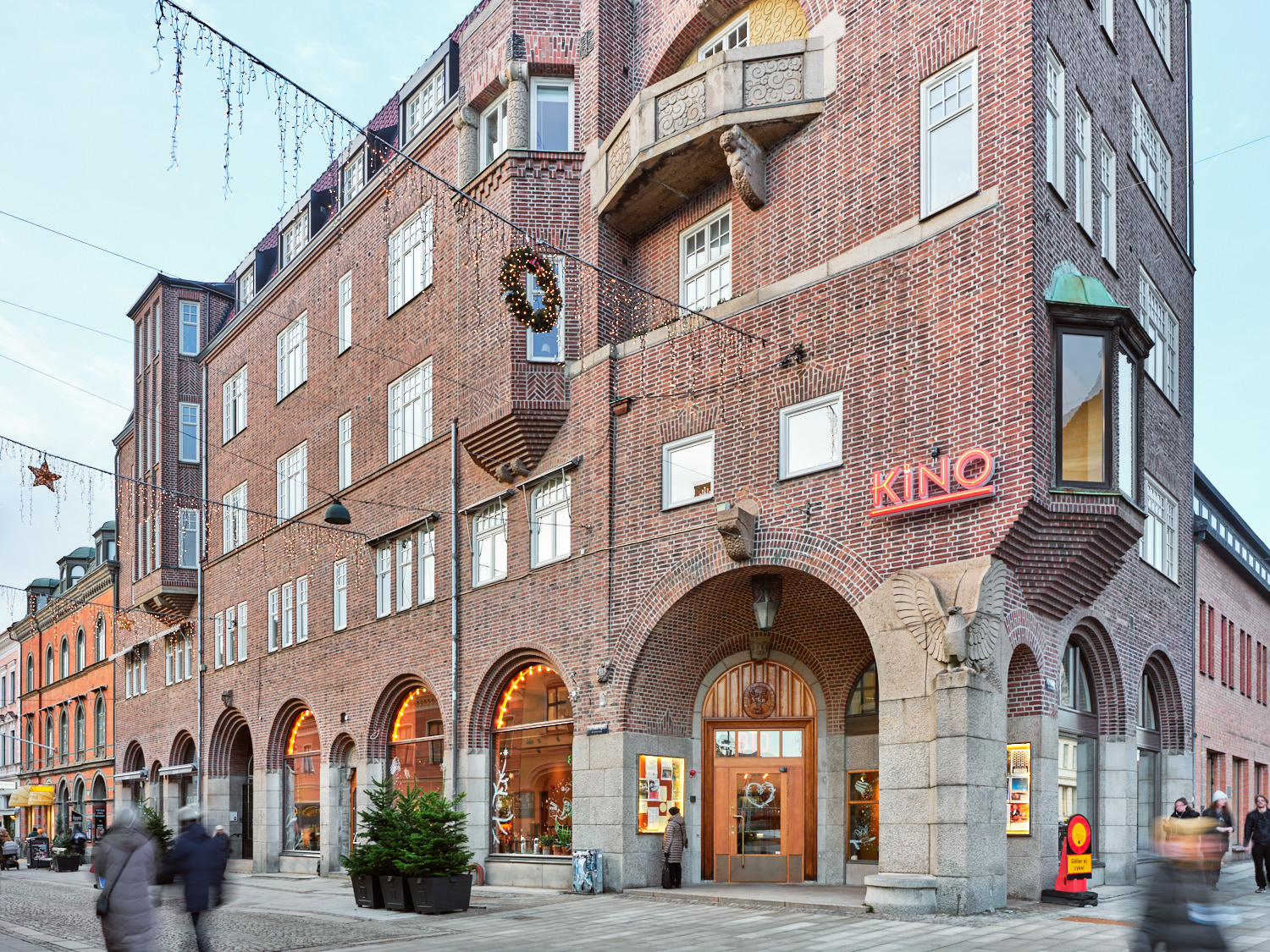 Gatubild av en äldre tegelbyggnad med välvda entréer, skyltfönster och en upplyst biografskylt med texten Kino.