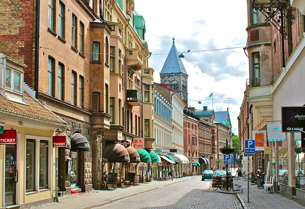 Stadsgata i centrala Lund med äldre byggnader i olika stilar längs båda sidor. Flera butiker och restauranger med skyltar och markiser syns i gatubilden. Längst bort i bakgrunden reser sig Lunds domkyrka med sina karakteristiska torn. Några bilar är parkerade längs gatan och en cykel står lutad mot en stolpe