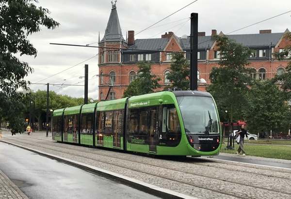 Grön spårvagn från Skånetrafiken på spårvägen i Lund, Sverige. Spårvagnen kör längs en gata med kullerstenar, omgiven av träd och ett stort rött tegelhus med torn i bakgrunden. Himlen är mulen och en person syns gå över gatan till höger.