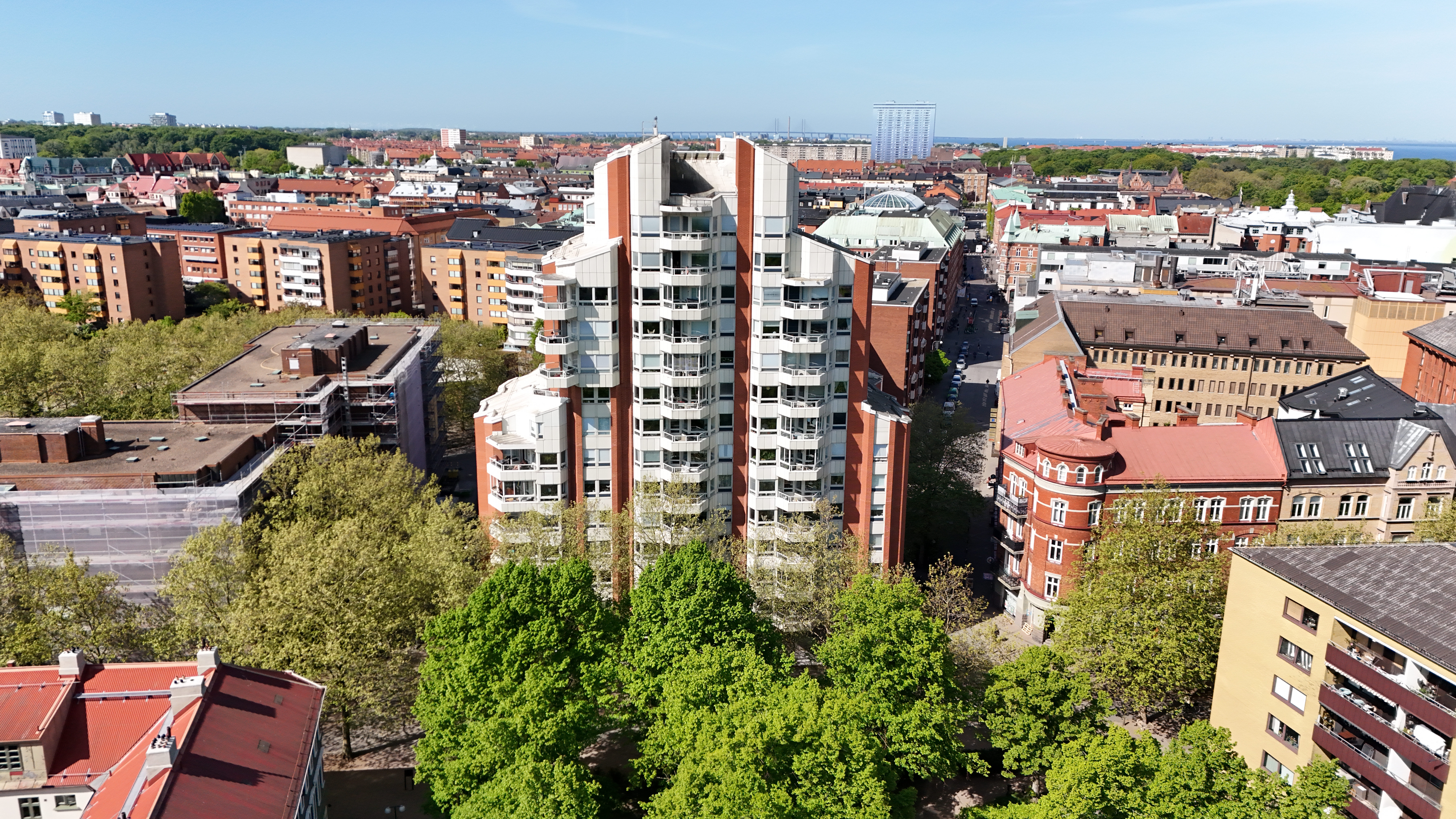 Flygbild över ett stadskvarter med bostadshus, grönskande träd och en hög vit byggnad i centrum.