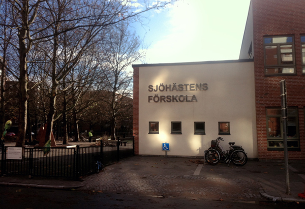 Entrén till Sjöhästens förskola med en vit fasad där namnet står skrivet i stora bokstäver. Två cyklar står parkerade framför byggnaden, och till vänster syns en lekplats med staket runt och träd som tappat sina löv. Himlen är delvis molnig med ett ljusinsläpp från solen.