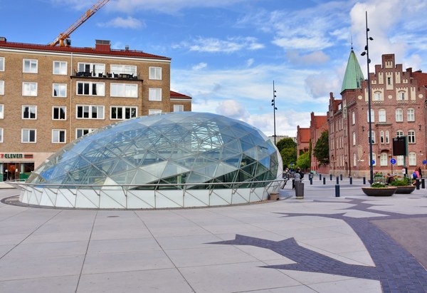 Triangeln i Malmö med den glasbeklädda kupolen som utgör en av entréerna till stationen. I bakgrunden syns både äldre byggnader i tegel och moderna flerbostadshus. Torget är öppet och belagt med ljusa plattor, och himlen är blå med några moln.