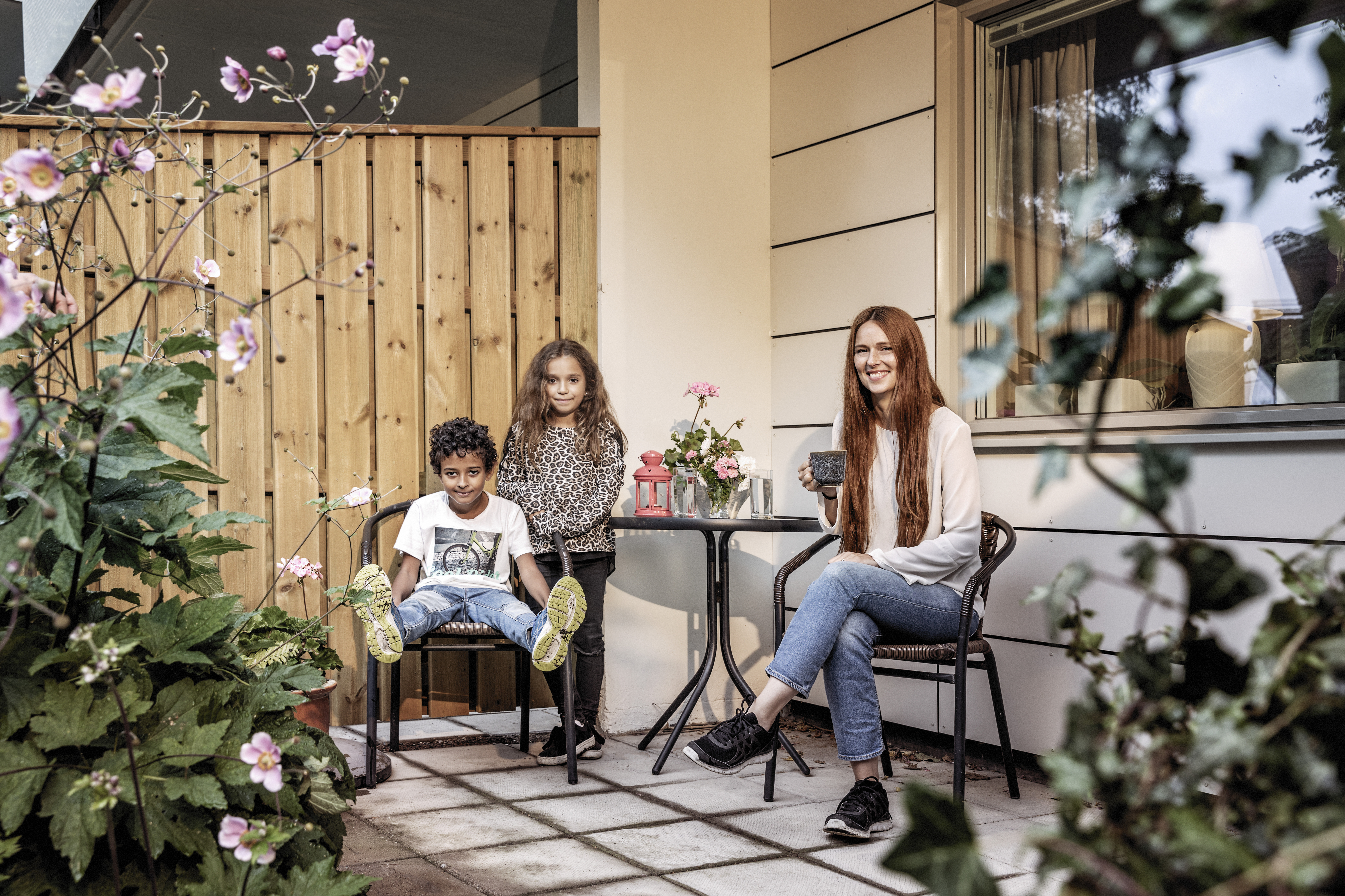 En kvinna och två barn sitter vid ett litet bord på en uteplats omgiven av blommor och grönska.