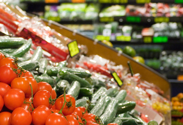 En välfylld frukt- och grönsaksavdelning i en livsmedelsbutik. I förgrunden syns klasar av röda tomater och prydligt staplade gurkor. I bakgrunden finns fler hyllor med grönsaker och frukter i olika färger, samt prisskyltar med gula och gröna etiketter.