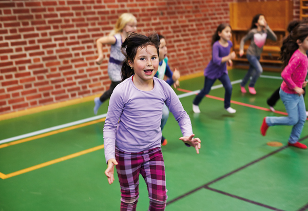 En grupp barn springer och leker i en gymnastiksal med grönt golv och röd tegelvägg. I förgrunden syns ett barn i lila tröja och rutiga byxor springa mot kameran med ett glatt uttryck. Flera andra barn i färgglada kläder syns i bakgrunden i rörelse, vilket skapar en livlig och energifylld stämning.
