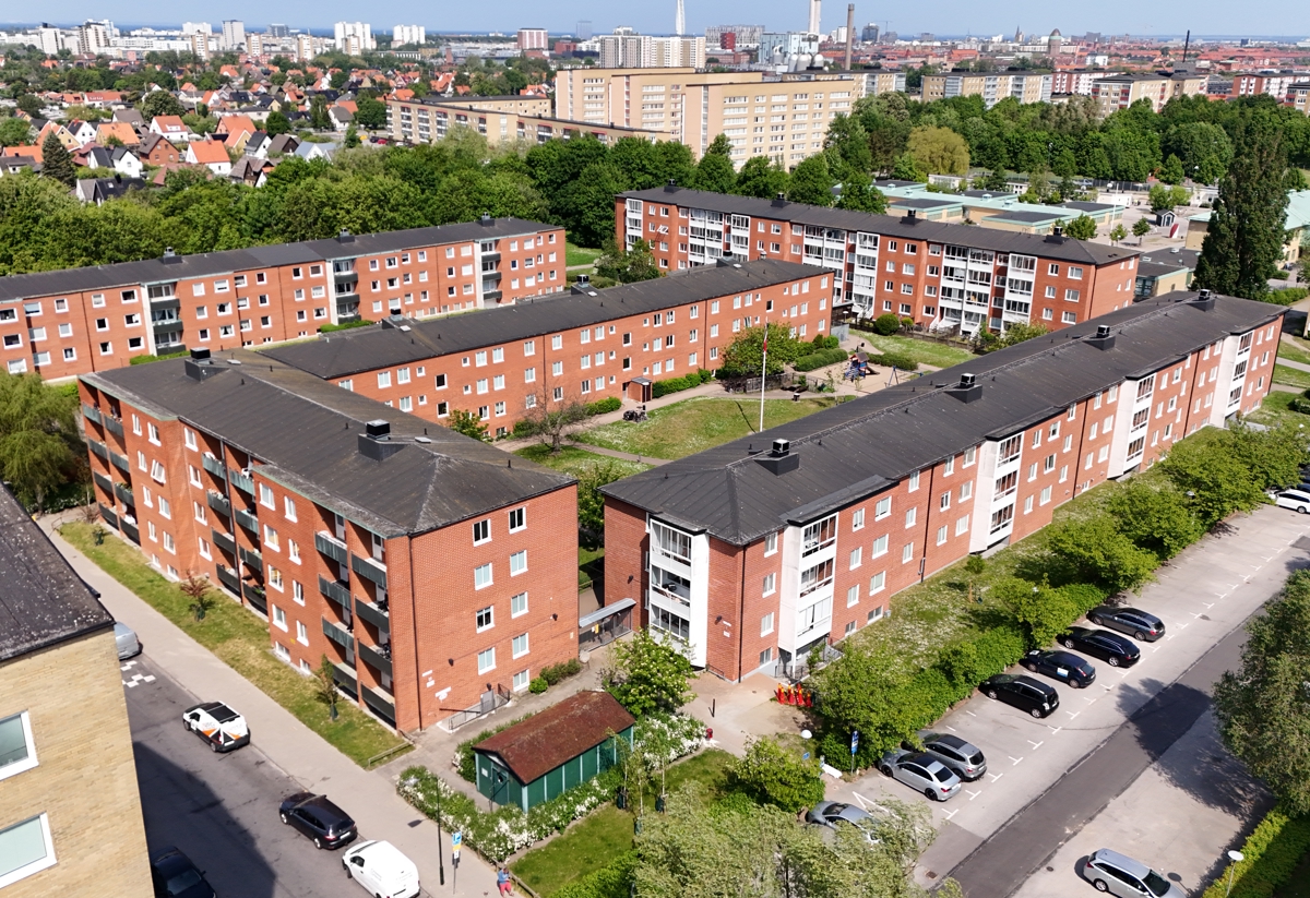 Flygbild över ett bostadsområde med flera röda flerbostadshus, innergårdar, grönska och parkerade bilar.