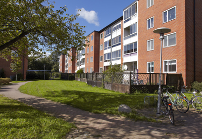 Ett bostadsområde med fyravånings flerfamiljshus i rött tegel, med inglasade balkonger. I förgrunden syns en grön innergård med grusgång, cyklar parkerade vid en lyktstolpe och ett träd som skuggar gången. Soligt väder och klarblå himmel förstärker en trivsam och lugn atmosfär.