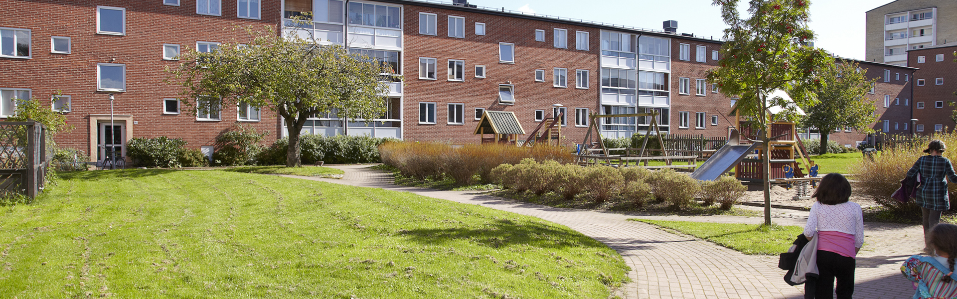 Ett bostadsområde med låga tegelhus och en grön innergård med lekplats. I förgrunden leder en röd gångväg genom gräset där en vuxen och två barn är på väg mot lekplatsen. Solen skiner från en klarblå himmel och ger hela området en ljus och inbjudande känsla.