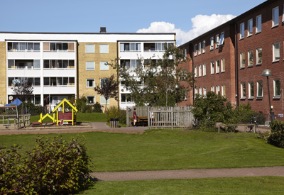 Ett bostadsområde med röda och gula flerfamiljshus i tre till fyra våningar. Mellan husen finns en grön gård med lekplats, grusgångar och planteringar. En färgglad lekställning i gult och rött syns i förgrunden och skapar en inbjudande miljö för barnfamiljer.