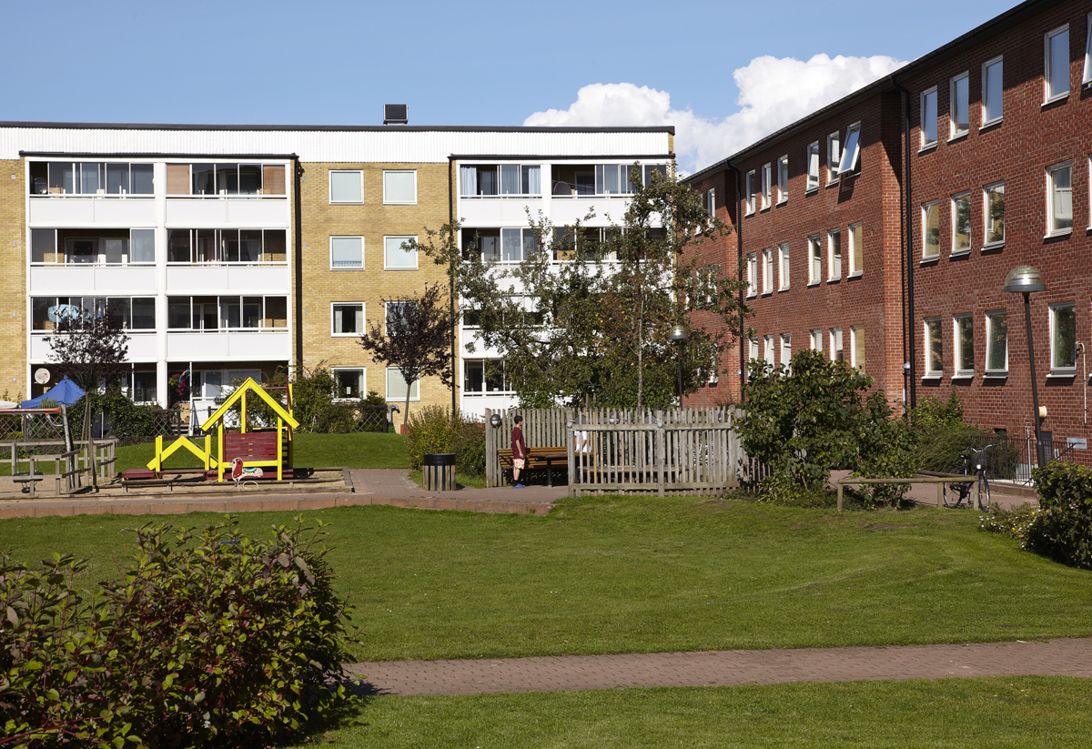 Ett bostadsområde med röda och gula flerfamiljshus i tre till fyra våningar. Mellan husen finns en grön gård med lekplats, grusgångar och planteringar. En färgglad lekställning i gult och rött syns i förgrunden och skapar en inbjudande miljö för barnfamiljer.