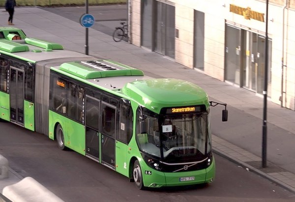 En grön ledbuss kör på en stadsgata i dagsljus. Den kör förbi ett modernt hus med stora fönster, och på trottoaren syns bland annat en cykel och ett par gångtrafikanter.