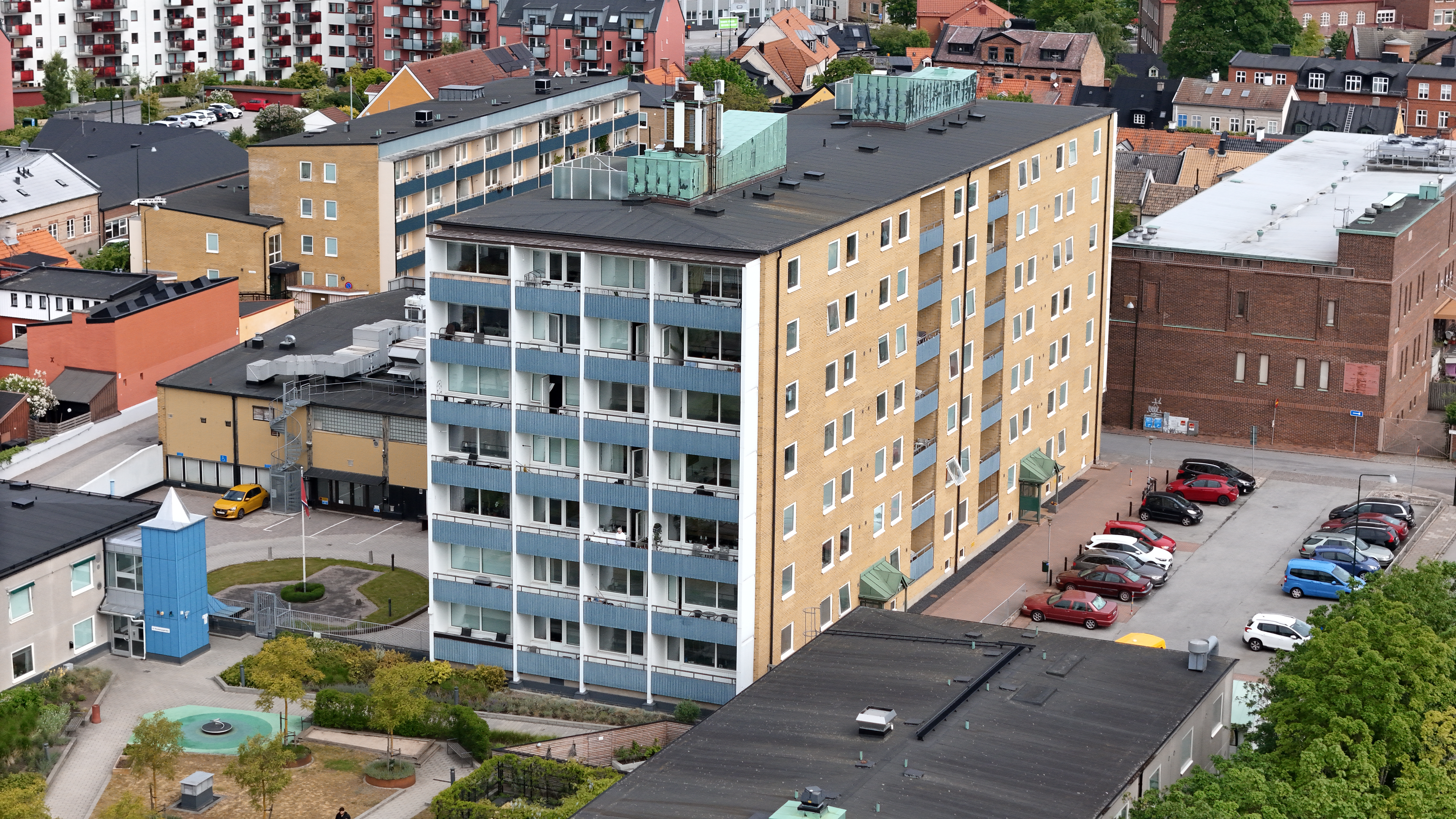 Flygbild över ett flerbostadshus med balkonger, omgivet av parkeringsplatser, lekplats och stadsmiljö.