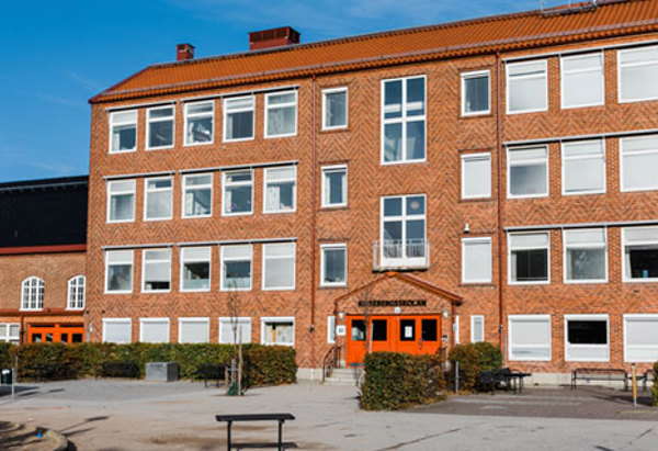 En skolbyggnad i tegel med flera våningar och ett rött tegeltak. Fönstren är symmetriskt placerade och många har vita persienner neddragna. I mitten finns en ingång med dubbeldörrar målade i orange. Framför skolan finns en öppen gårdsplan med bänkar, staket och några buskar. Himlen är klarblå.