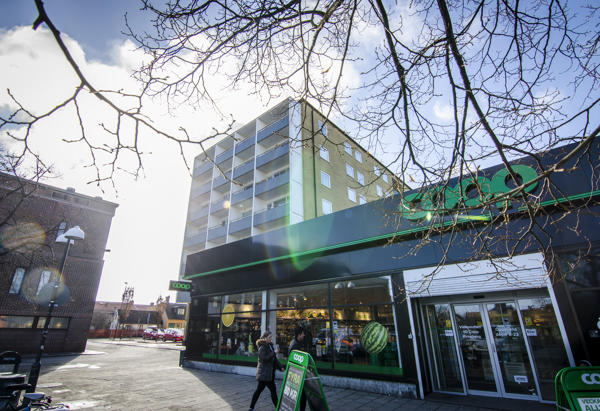 En Coop-butik i ett stadsområde, med ett flerfamiljshus i bakgrunden. Solen skiner genom trädgrenar utan löv, vilket skapar ett motljus med solreflexer i bilden. En person är på väg in eller ut genom butikens ingång, och gatuscenen visar en blandning av bostäder och kommersiella byggnader.