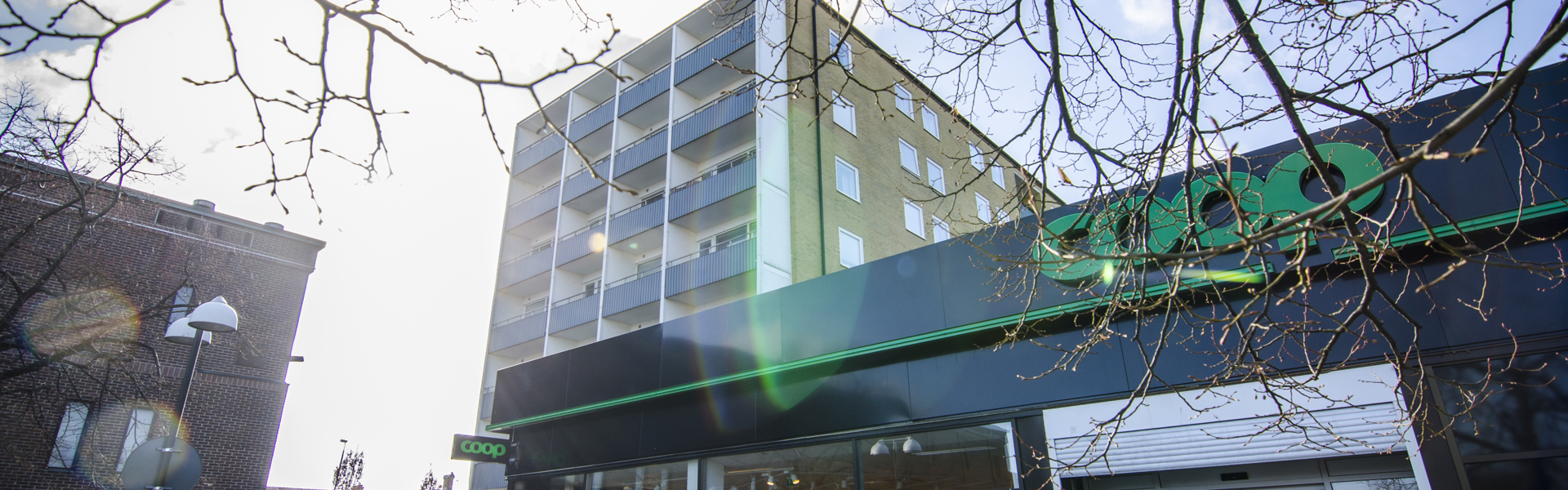 En Coop-butik i ett stadsområde, med ett flerfamiljshus i bakgrunden. Solen skiner genom trädgrenar utan löv, vilket skapar ett motljus med solreflexer i bilden. En person är på väg in eller ut genom butikens ingång, och gatuscenen visar en blandning av bostäder och kommersiella byggnader.