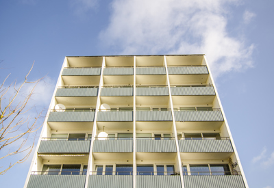 Ett flerbostadshus sett rakt framifrån underifrån, med åtta våningar och balkonger i rad. Byggnaden har vita balkongräcken med ett blågrönt inslag och symmetrisk arkitektur. Himlen är klarblå med några vita moln, vilket skapar en ljus och positiv atmosfär i bilden.