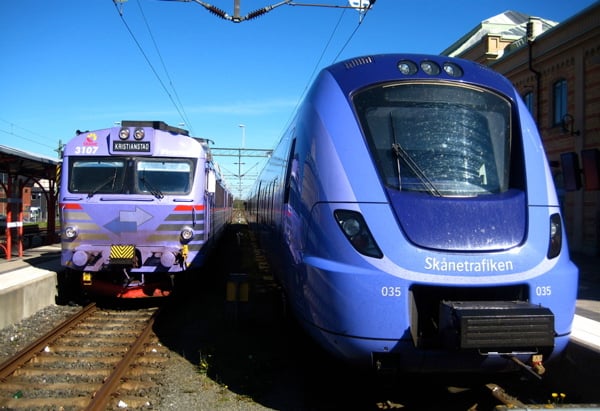 Två tåg står sida vid sida vid en perrong. Till vänster syns ett äldre, lila tåg med destinationen "Kristianstad". Till höger står ett modernt blått Pågatåg från Skånetrafiken med numret 035. Himlen är klarblå och en äldre stationsbyggnad syns i bakgrunden.