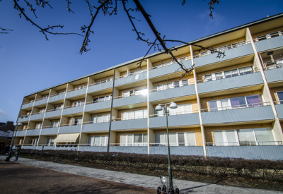Ett fyravånings flerfamiljshus med långa rader av balkonger, fotograferat snett framifrån under en klarblå himmel. Fasaden har blågrå balkongräcken och gula pelare som ramar in varje sektion. Framför huset finns en gångväg, en buskrad och en lyktstolpe. Grenar från ett träd syns upptill i bildens förgrund.