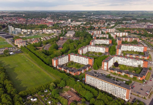 Flygfoto över ett bostadsområde med flera stora flerfamiljshus i rader, omgivna av grönska och träd. Till vänster syns stora gräsfält och idrottsplaner, och i bakgrunden breder staden ut sig mot horisonten. Bebyggelsen är tät med inslag av både moderna och äldre byggnader.
