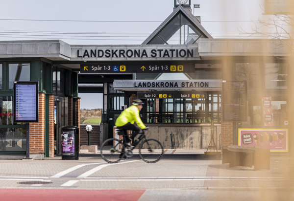 En person på cykel i gul reflexjacka passerar framför entrén till Landskrona station, med stationsskyltar och perrongangivelser tydligt synliga i bakgrunden.