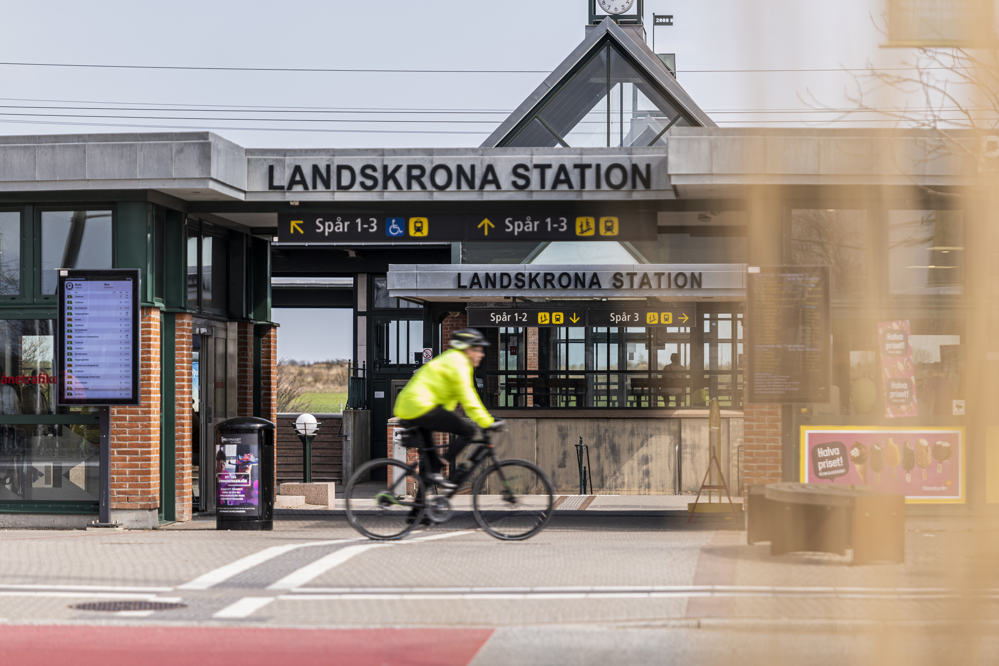 En person på cykel i gul reflexjacka passerar framför entrén till Landskrona station, med stationsskyltar och perrongangivelser tydligt synliga i bakgrunden.