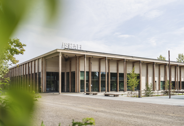 En modern, ljus byggnad (ishall) med stora fönster och träpelare omgiven av träd och grönska, sedd från sidan mot en ljus himmel.